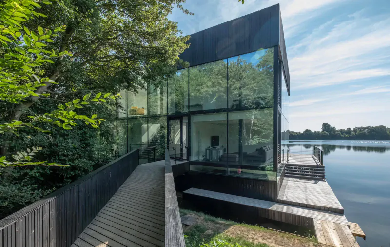 Modern angular glass house on a wooden dock by the lake, surrounded by green trees under a partly cloudy sky.