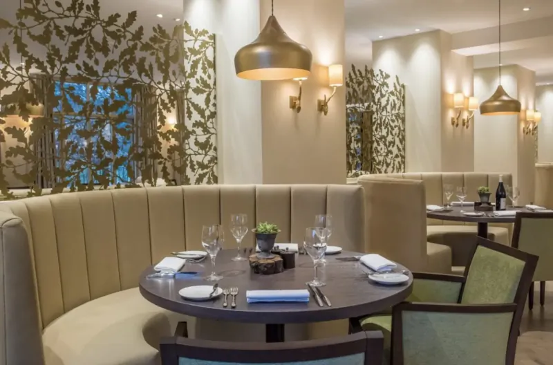 Elegant dining area at Careys Manor Hotel with curved beige banquette, round wooden table set with wine glasses, gold pendant lights, oak leaf wall screens.
