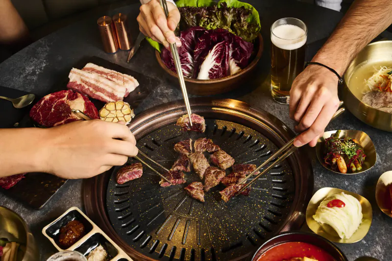 Hands using tongs to grill beef and pork on a Korean BBQ table with lettuce wraps, banchan, and beer.