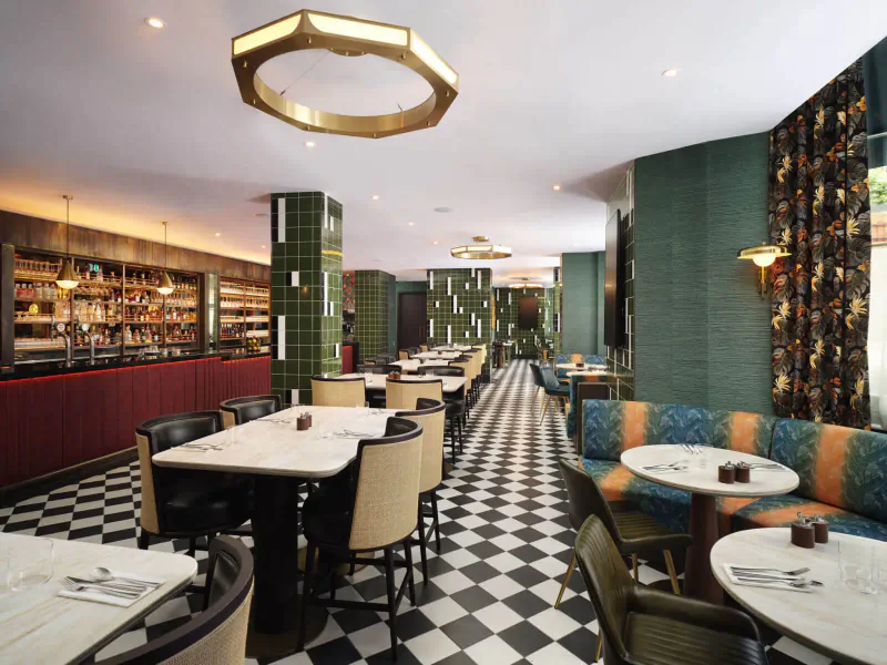 Elegant restaurant interior with Art Deco green tiles, gold lighting, checkered floors, wooden tables, and velvet seating.