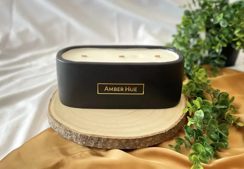 Black 'Amber Hue' candle with amber flecks on wood slice, surrounded by green plants on gold fabric.