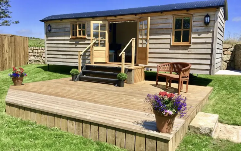 Wooden luxury holiday cottage with blue roof on deck, steps, bench, potted flowers, surrounded by green lawn and fence.