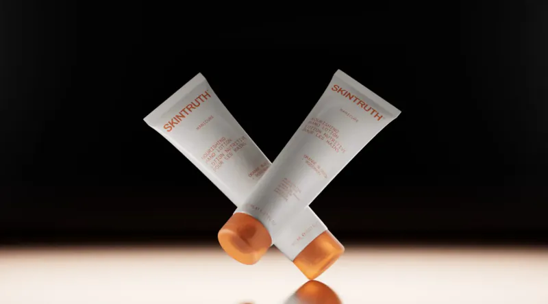 Two crossed Skintruth facial care tubes with orange caps on reflective surface, black background.