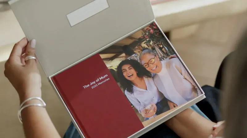 Woman opening gray box revealing red 'The Joy of Aging' photo book of smiling woman and older lady.