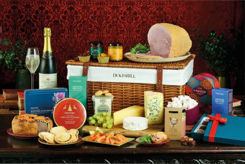Dukeshill luxury Christmas hamper with champagne, glazed ham, cheeses, biscuits, and festive treats on a wooden table.