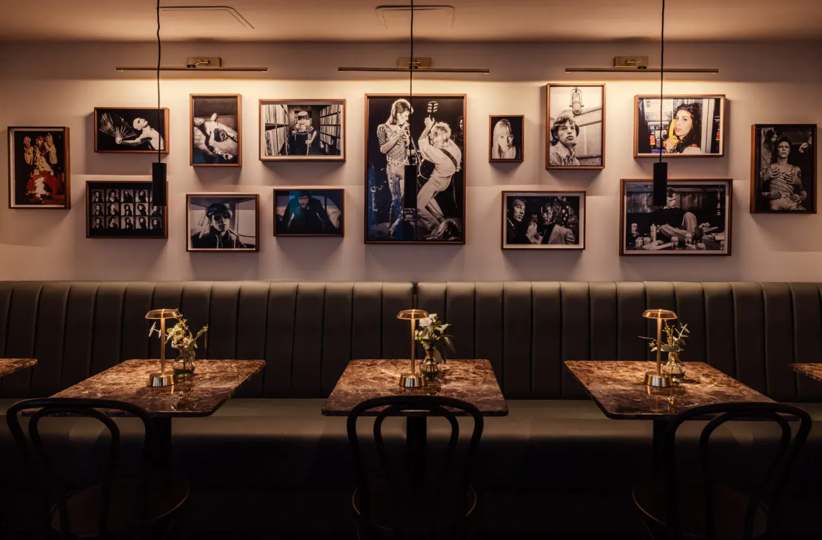 Elegant restaurant interior with black-and-white vintage photos on wall, tufted bench seating, round tables with lamps and flowers
