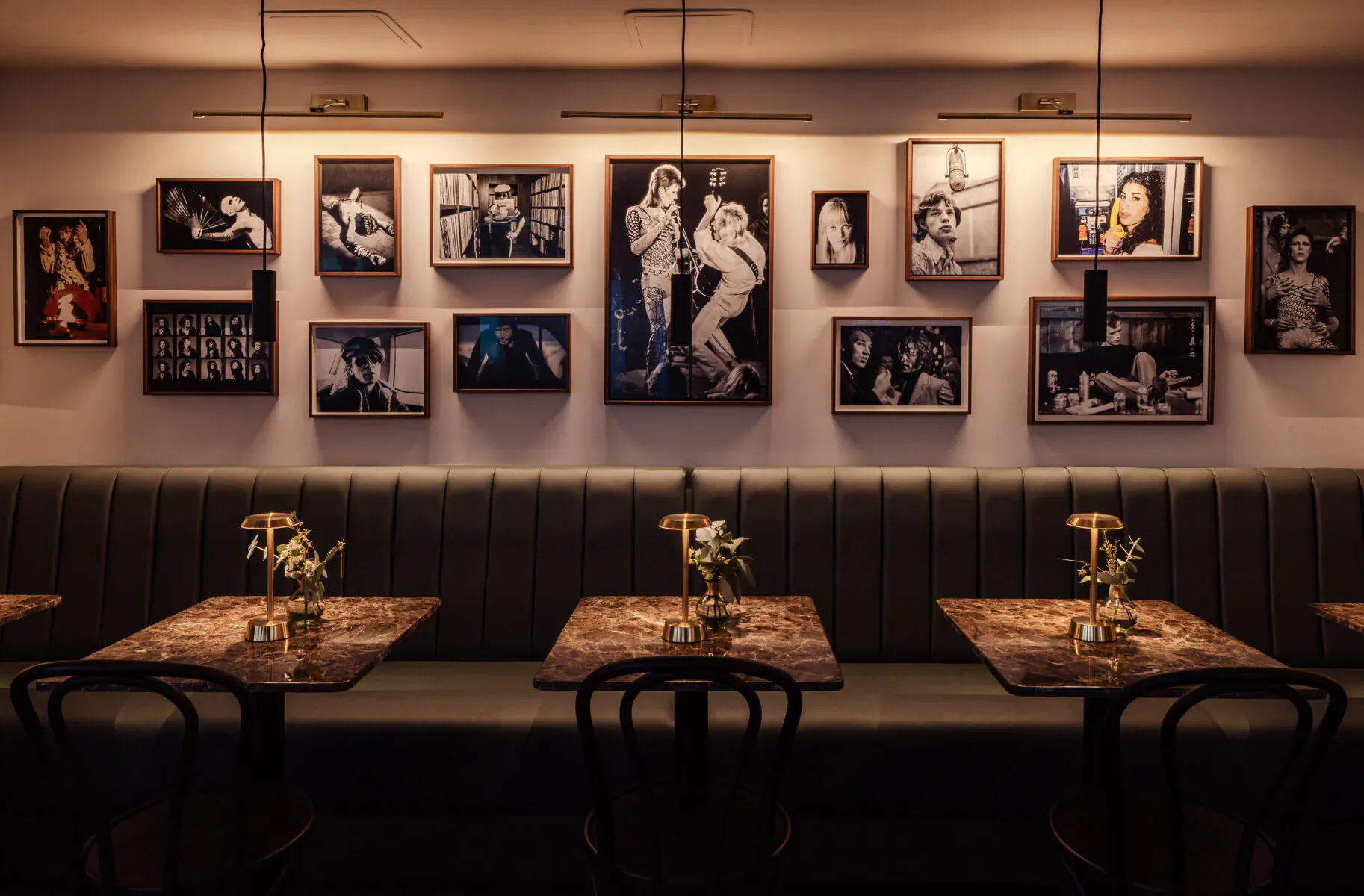 Elegant restaurant interior with black-and-white vintage photos on wall, tufted bench seating, round tables with lamps and flowers