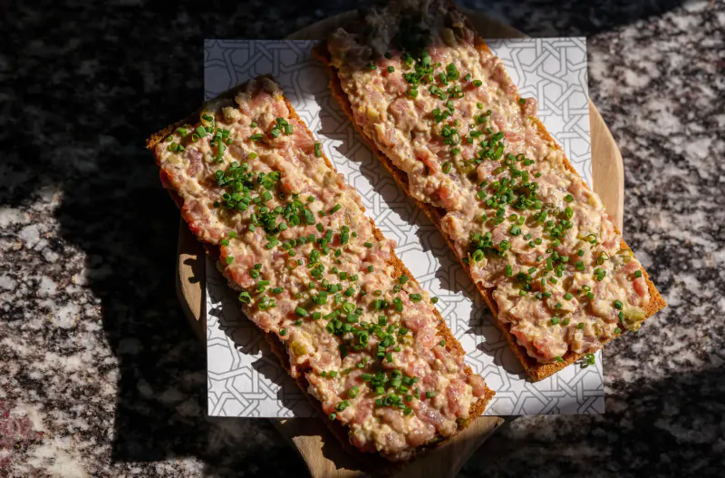 Two garnished pink meat tartines topped with chopped green herbs on white paper atop wooden board