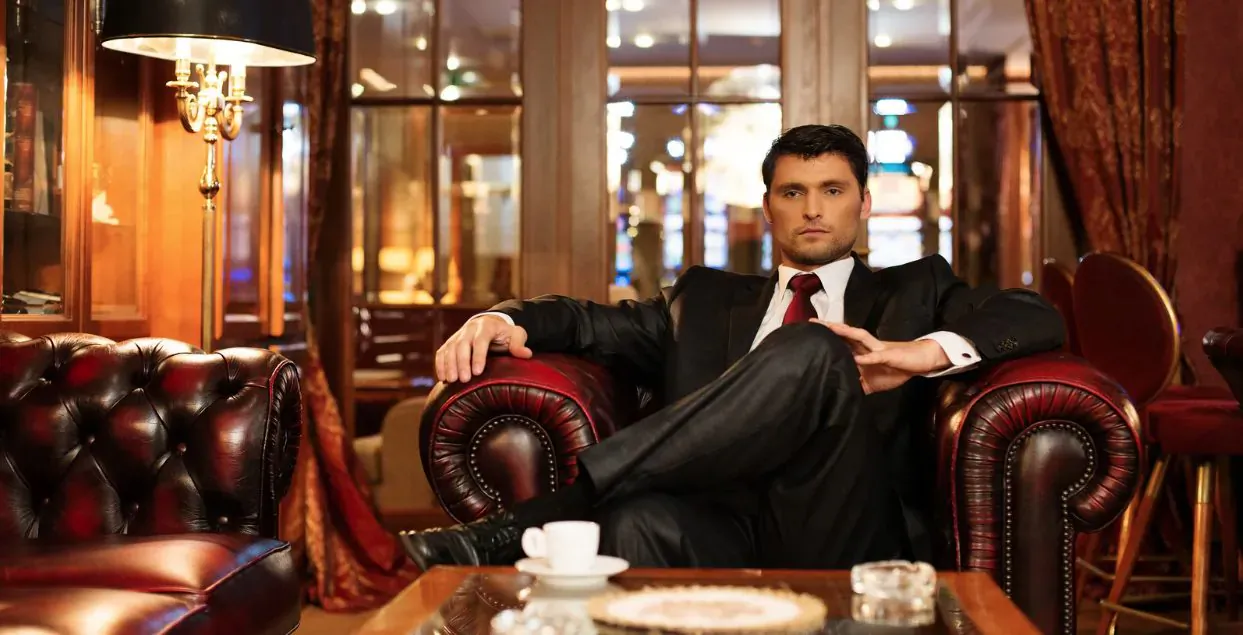 Confident man in suit and red tie sits relaxed in red leather armchair in luxurious room with coffee cup on table