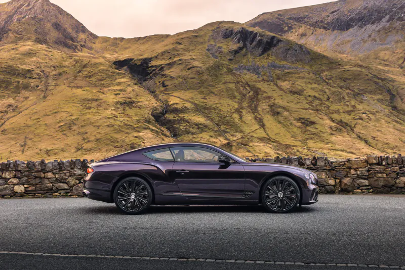 Purple Bentley GT Mulliner Blackline coupe parked on mountain road with green hills and stone wall at sunset.