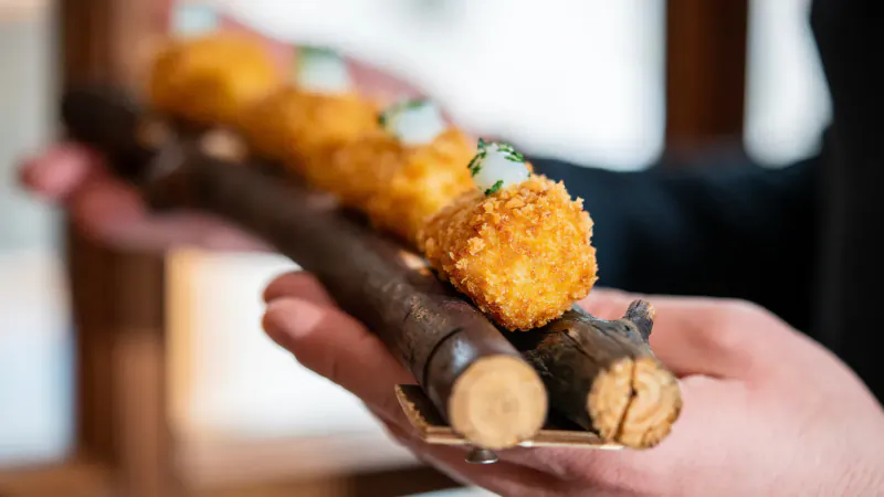 Chef's hands holding a wooden branch skewer with golden arancini topped with green herbs at El Restaurant El Llorenç