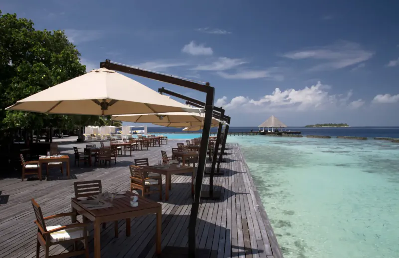 Wooden deck at Coco Bodu Hithi resort with beige umbrellas over tables, overlooking turquoise lagoon and distant island.