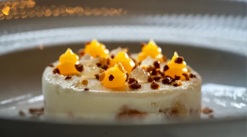 Round white dessert topped with golden mango pieces and granules on a plate