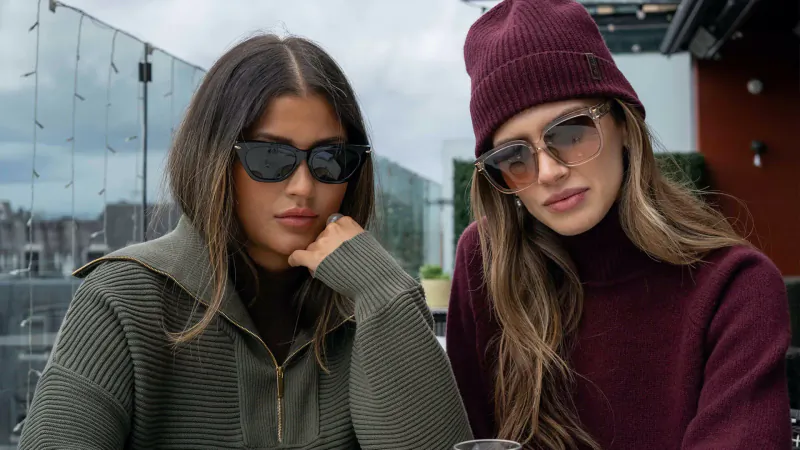Two stylish women in sunglasses and knitwear—one in green hoodie, one in maroon turtleneck and burgundy beanie—pose on rooftop.