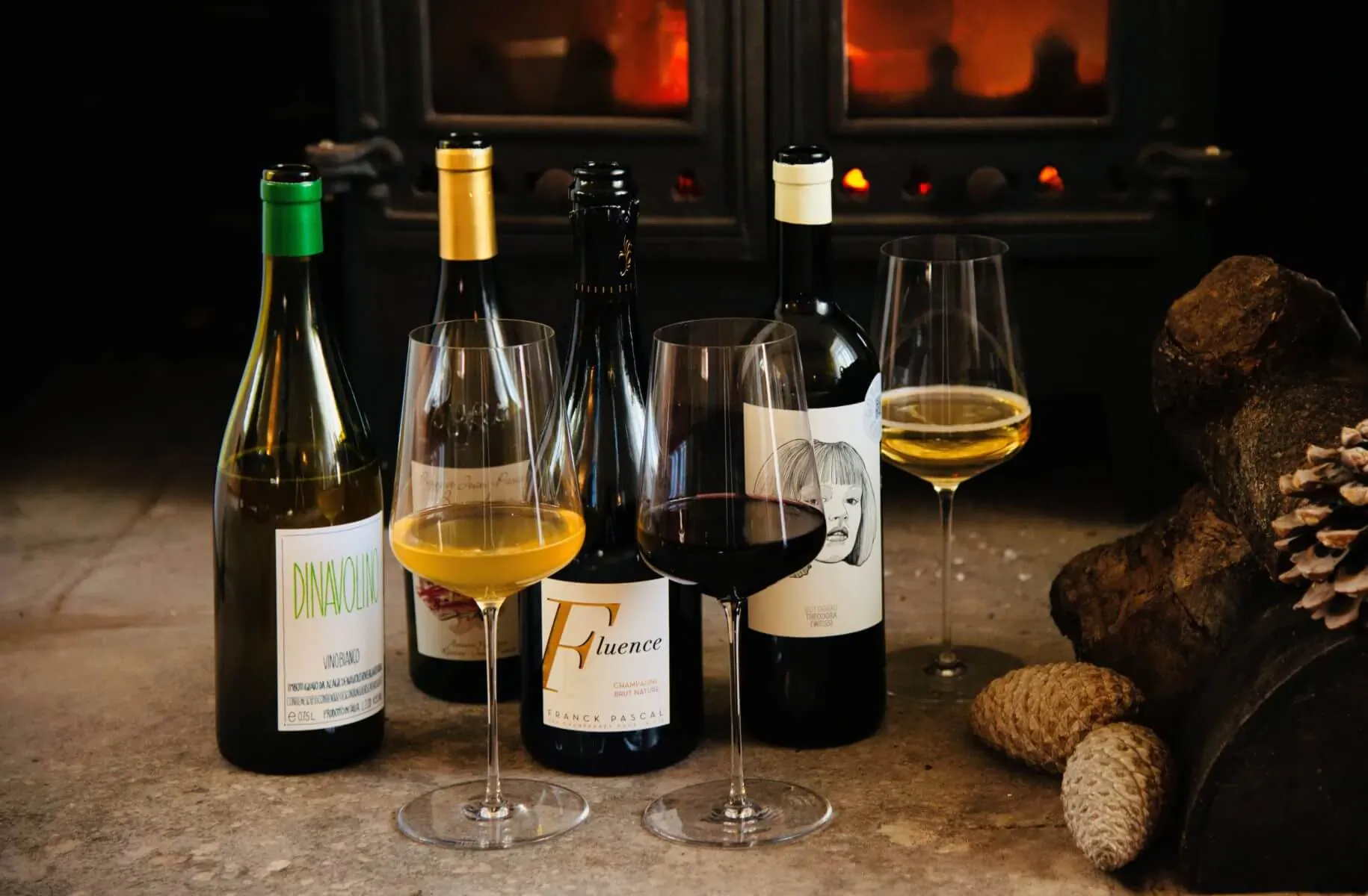 Assortment of natural wines in green, gold, and dark bottles with white and red pours beside a cozy fireplace.