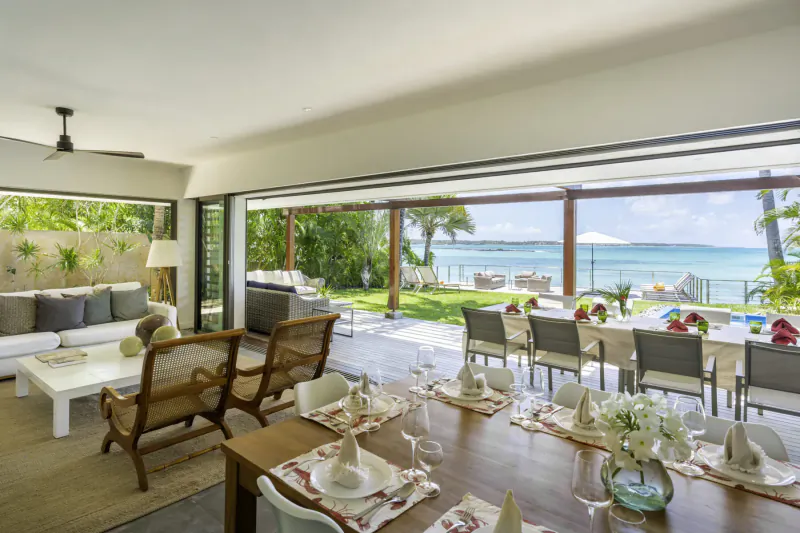 Spacious open-plan villa interior with wooden dining table set for meal, lounge seating, opening to lush garden, pool, and ocean view at Belle Mare beach.