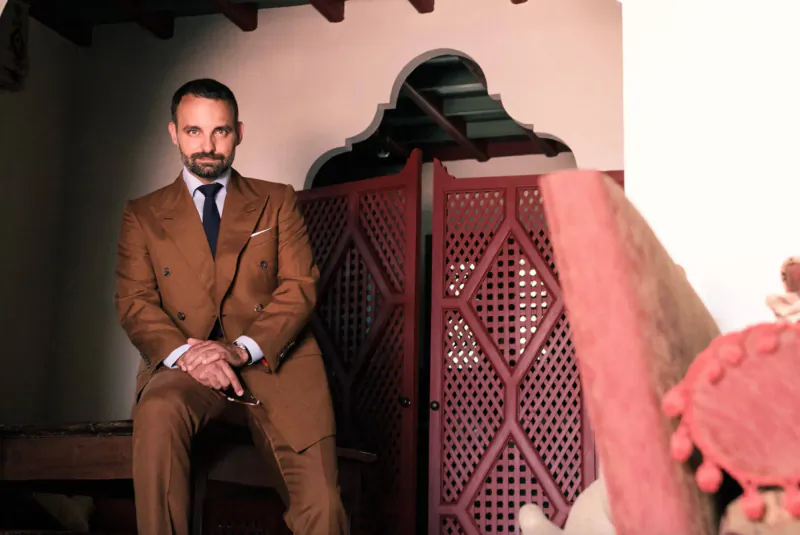 Man in tan Savile Row suit sits confidently by red latticed screen in Marbella gallery archway