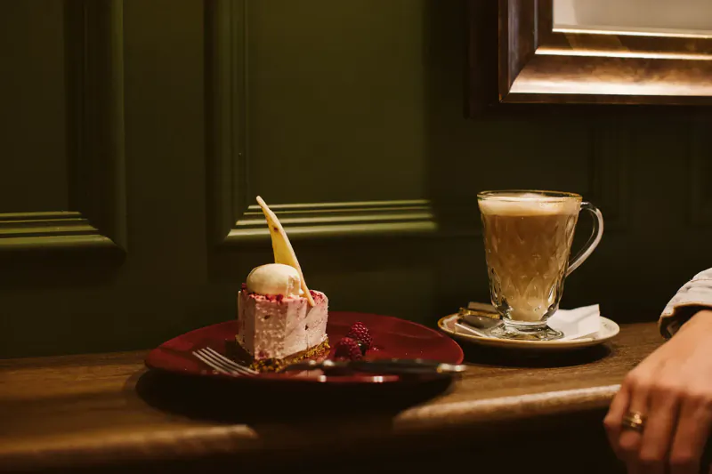 Close-up of pink cheesecake topped with ice cream and cream on red plate, beside frothy coffee cup on wooden table at The Rabbit Hotel.