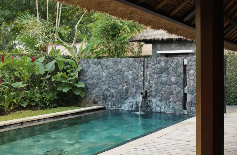Tropical Bali villa infinity pool with stone waterfall, lush greenery, thatched roof, overlooking Ayung River.