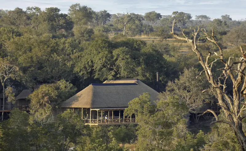 Thatched-roof lodge with veranda nestled in lush African savanna bushveld amid trees and hills.