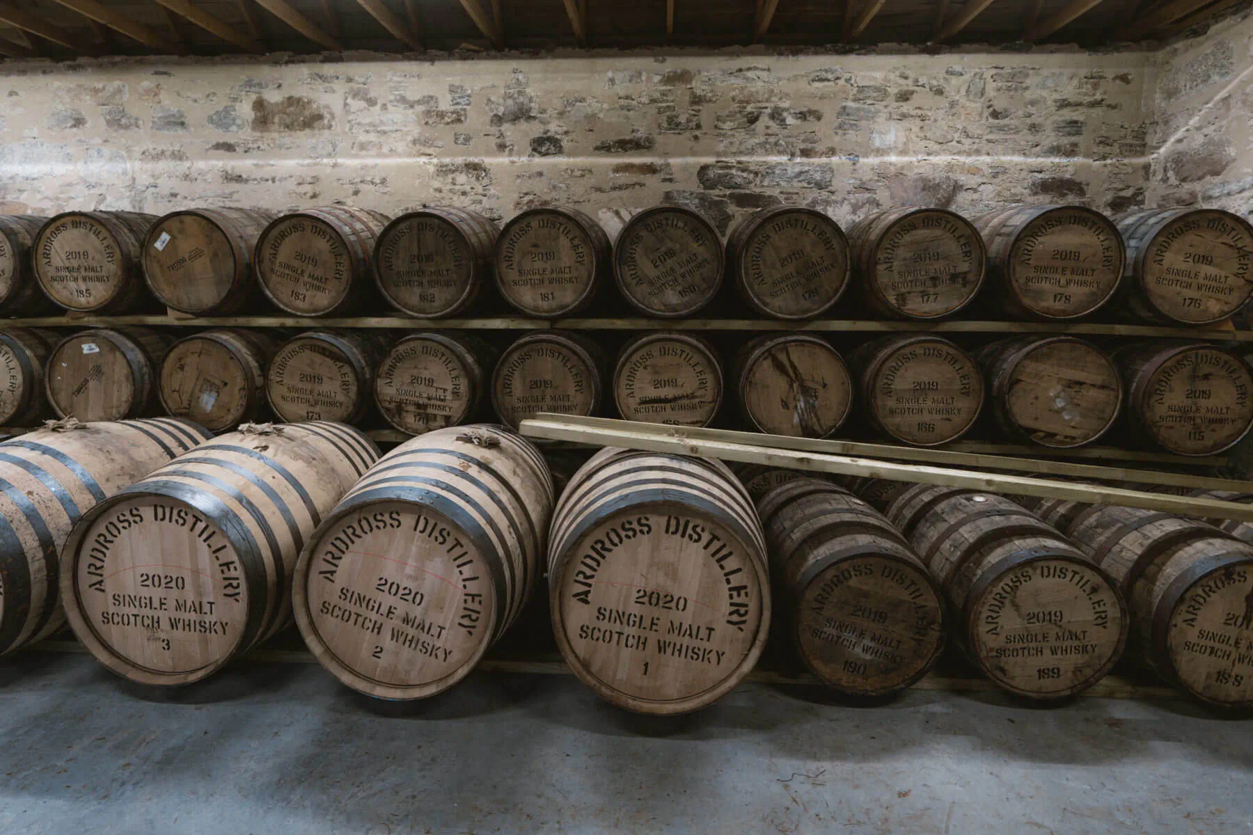 Rows of wooden whiskey barrels stamped 'Ardross Whisky Distillery 1980' stacked in rustic brick barrel storehouse.