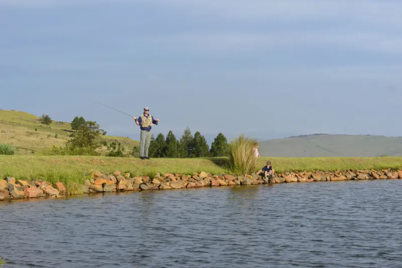 Two anglers fishing from grassy bank of serene pond amid rolling hills under blue sky.