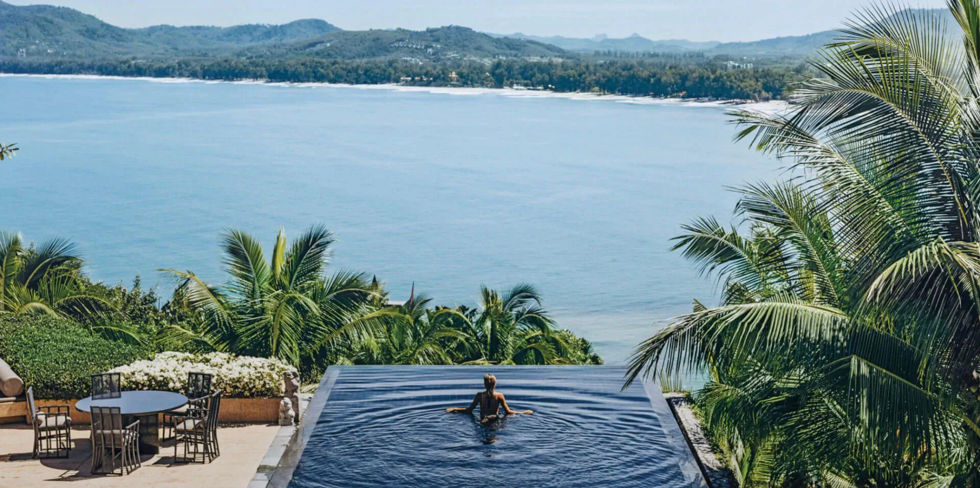 Woman swimming in infinity pool overlooking turquoise sea, palm trees, and distant mountains at luxury resort.