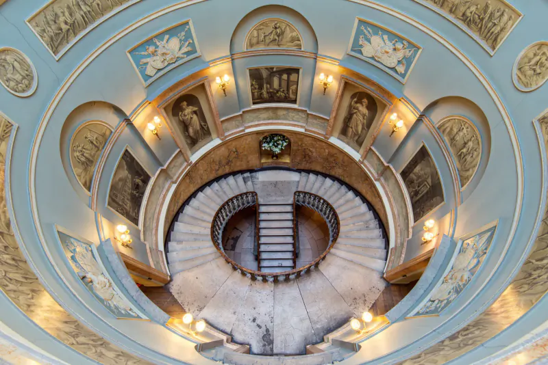 Aerial view of ornate circular staircase in Home House club, with blue domed ceiling, gilded frescoes, and wall lamps.