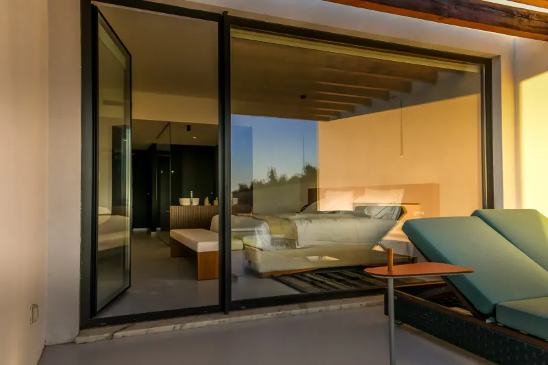 Open glass doors from modern bedroom to terrace with two teal sun loungers, sunset glow over landscape.