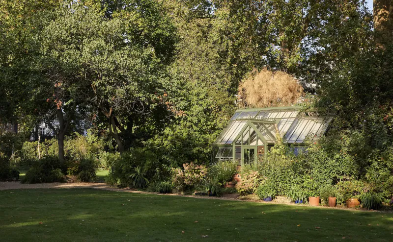 Glass greenhouse surrounded by lush green trees, plants, and terracotta pots in a grassy garden