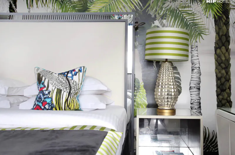 Luxury hotel bed with colorful patterned pillows, green-striped lamp on nightstand, palm leaf wallpaper backdrop