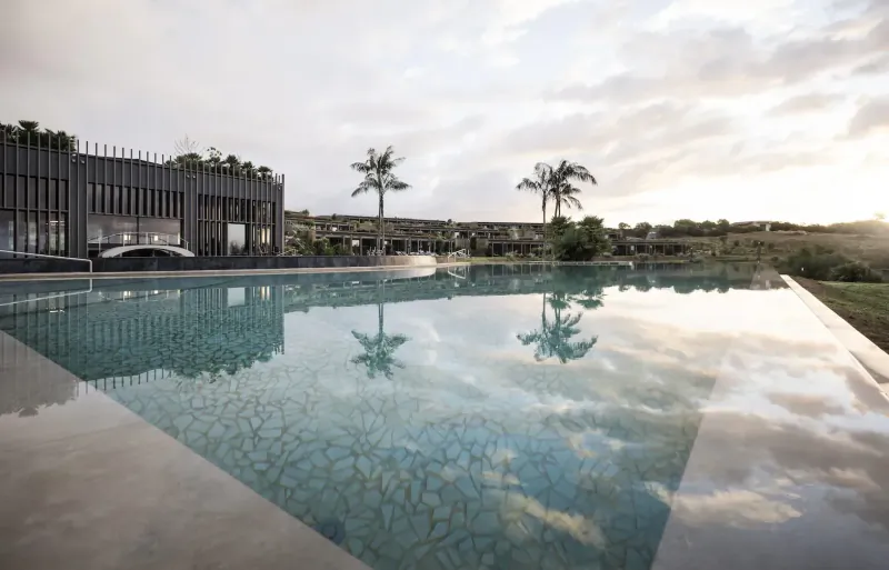 Infinity pool reflecting modern black lodge, palm trees, and sunset sky at ADLER Spa Resort Sicily.