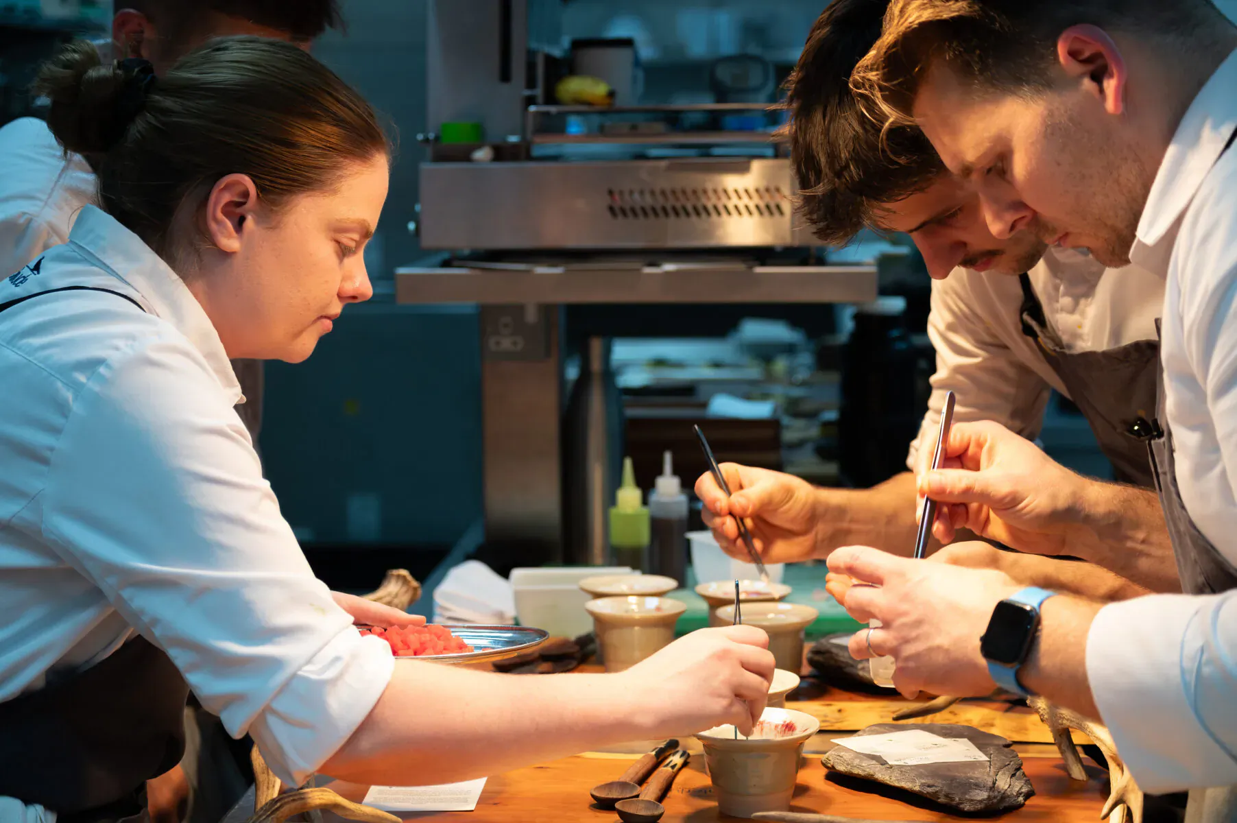 Chefs in white uniforms meticulously plating dishes with tweezers on a wooden counter in a professional kitchen.