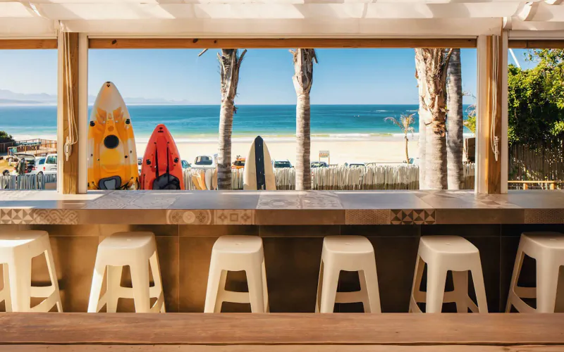 Seaside bungalow bar with surfboards leaning on counter, white stools, ocean and palm trees view through windows in Plettenberg Bay
