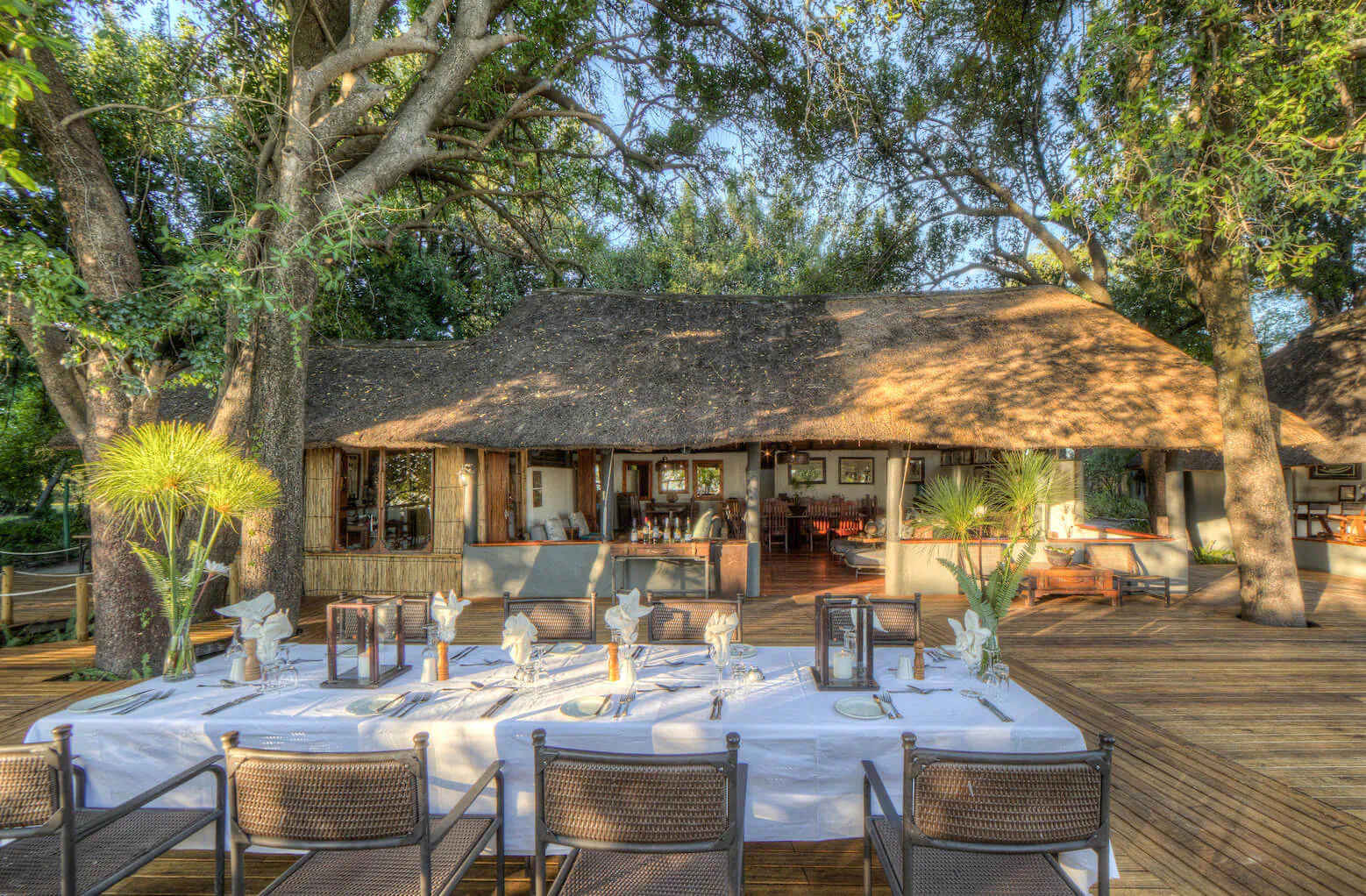 Elegant outdoor dining table set on wooden deck at Xugana Island Lodge, thatched building with trees and plants in background.