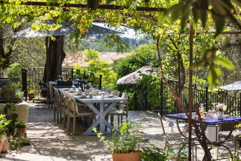 Outdoor terrace at Lou Calen in Provence with long white table set for dining under grapevines and umbrellas, lush garden view.