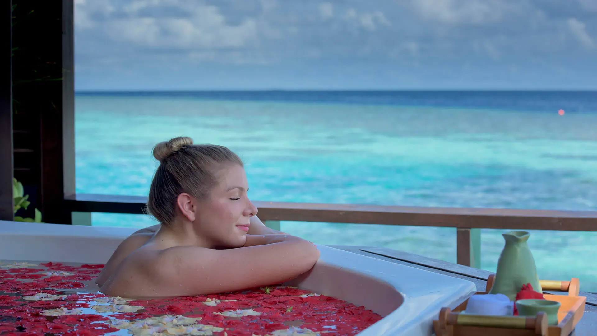 Blonde woman relaxing with eyes closed in red petal-filled hot tub on overwater villa deck with turquoise ocean view