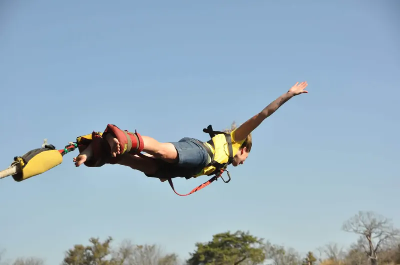 Person bungee jumping over Victoria Falls, mid-air dive in yellow harness, red shoes, blue sky and trees below.