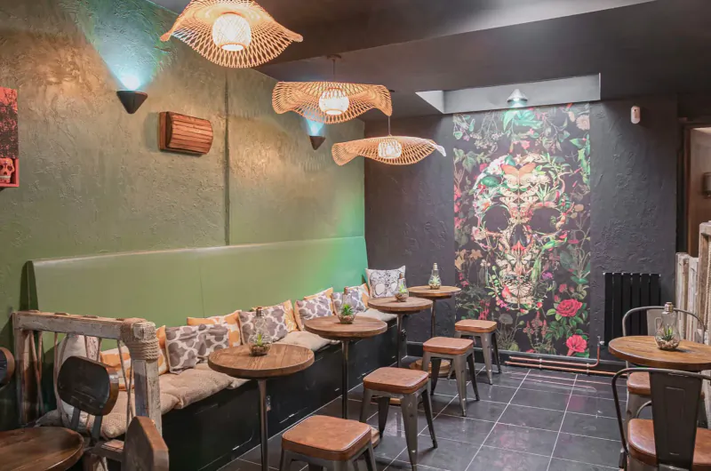 Cozy Mexican bar interior with green banquette, wooden stools, rattan lamps, and large floral skull wall mural.