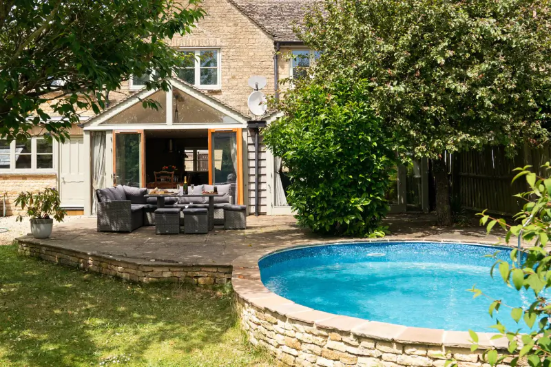 Cotswolds country house backyard with conservatory, outdoor seating on deck, and round blue pool amid lush gardens.