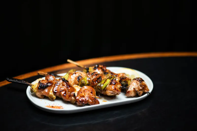 Grilled chicken and green pepper yakitori skewers on a white oval plate on a dark wooden table.