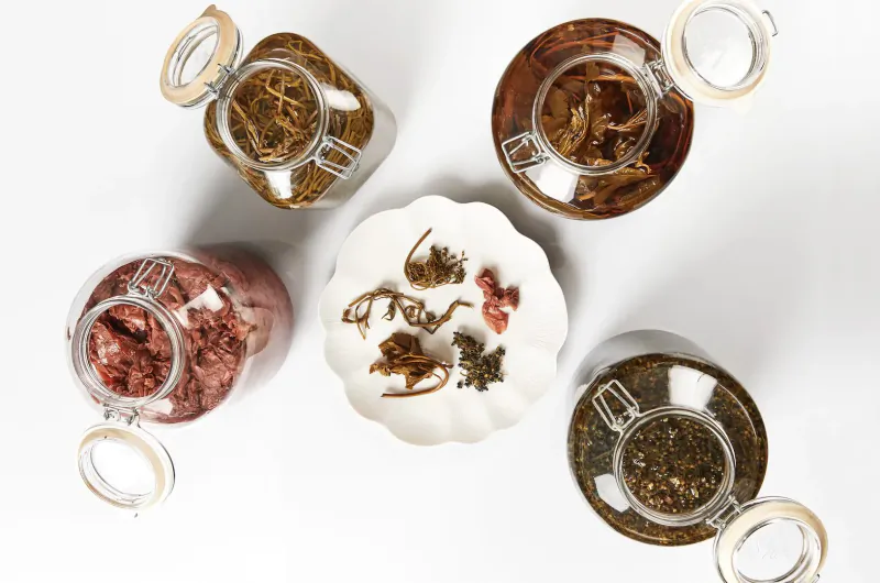 Overhead view of glass jars with dried herbs and pink seaweed on white plate, Scandinavian style.