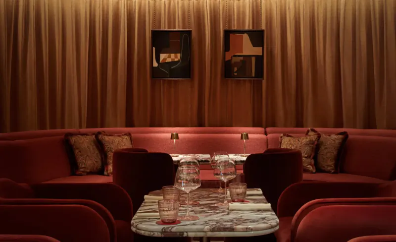 Revery Bar’s elegant red velvet booth seating with marble table, wine glasses, abstract art, and warm drapes for afternoon tea.