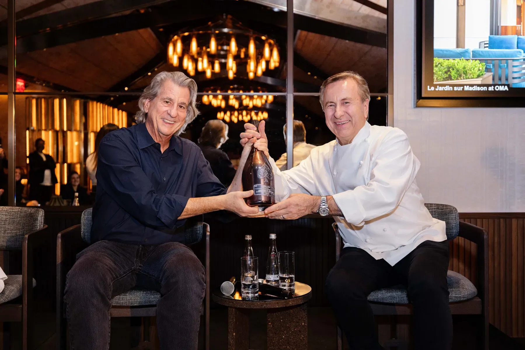Two men in suits and chef whites toasting with Armand de Brignac champagne bottle in elegant New York restaurant.