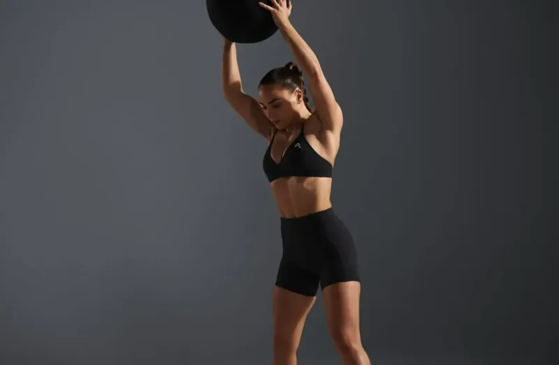 Fit woman with ponytail lifts heavy black medicine ball overhead in black sports bra and shorts, gym setting