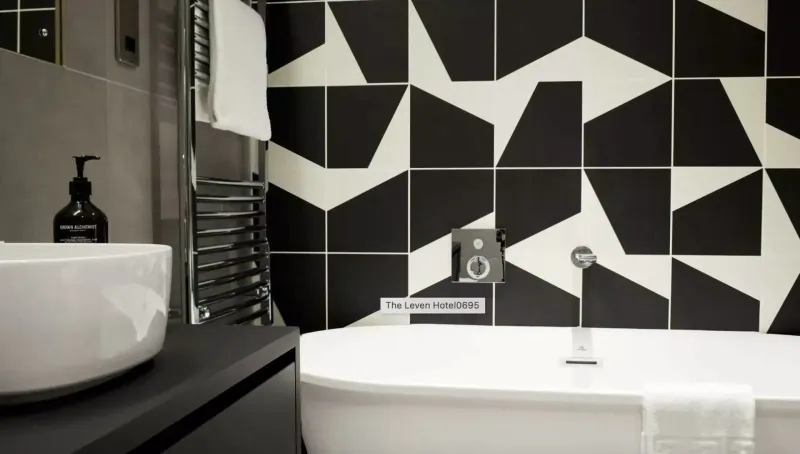 Modern bathroom with black and white geometric tiled wall, freestanding white tub draped with towel, black sink, ladder radiator, and soap dispenser.