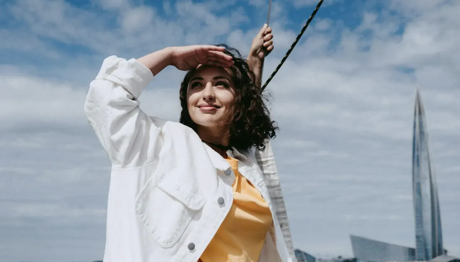 Smiling woman in white jacket and yellow top shades eyes on sailboat with tall sail and modern tower backdrop
