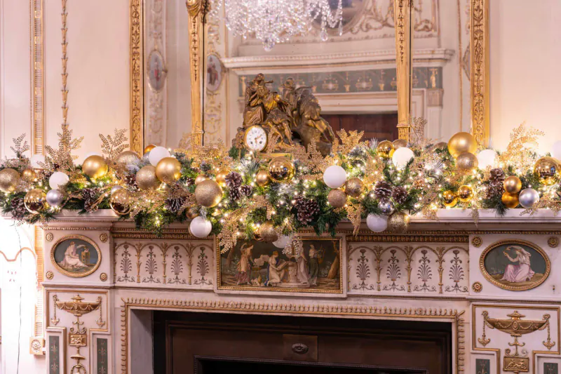 Opulent gold and white Christmas garland with lights, ornaments, and pinecones on ornate fireplace mantel at Home House, London.
