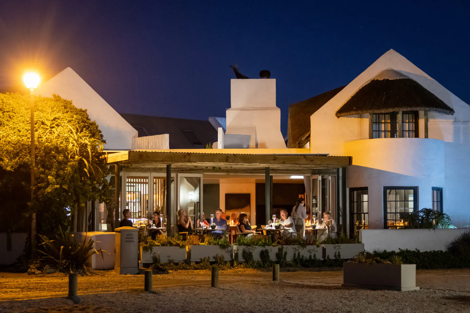 Evening exterior of Abalone Hotel's white Oyster Suites lounge with guests dining on terrace under streetlamp, coastal night.
