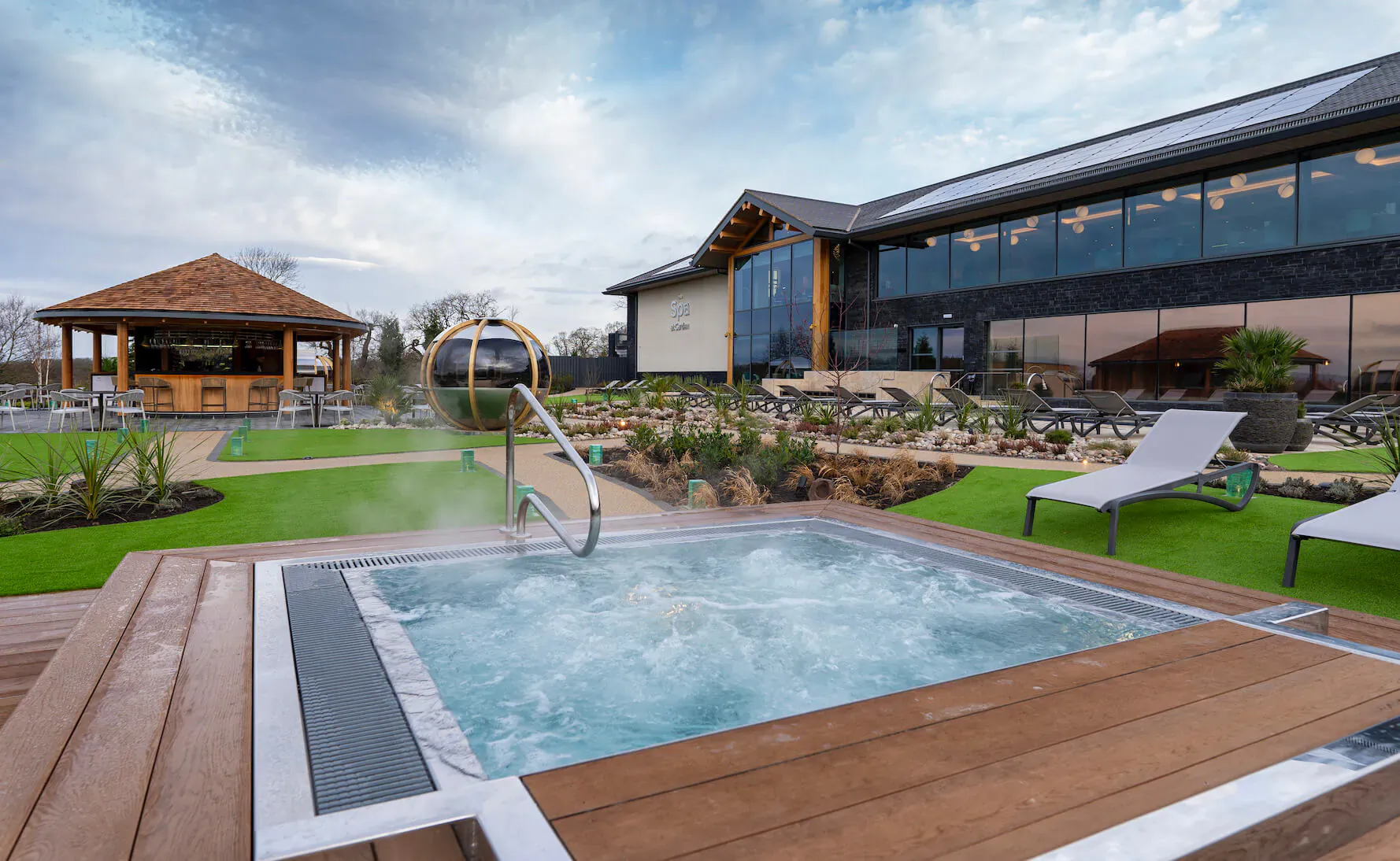 Bubbling hot tub in spa garden at Carden Park, with wooden pergola, loungers, and modern building under cloudy sky.
